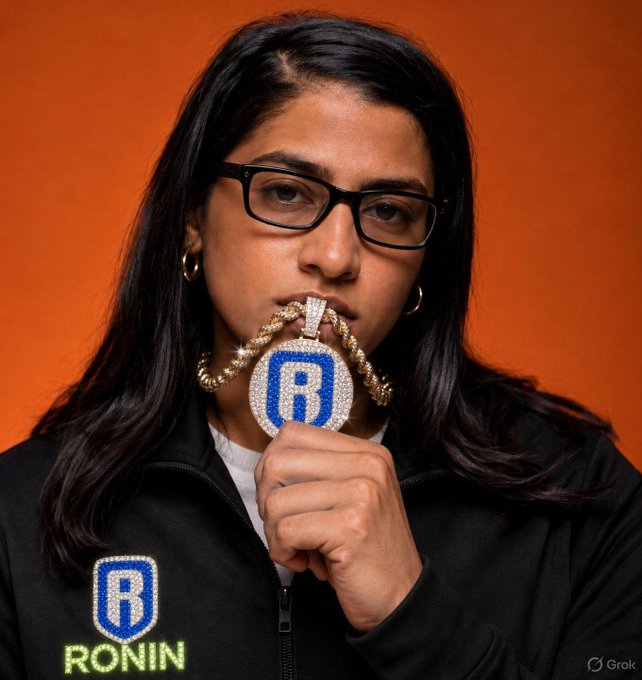 A portrait of a young woman with glasses holding a large diamond-encrusted 'R' pendant to her face; her black jacket features a 'RONIN' logo with an 'R', and the image has a Grok watermark.