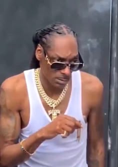 A man with braided hair wearing sunglasses and a white tank top looks down at his hands, accessorized with thick gold chains and a cross pendant.