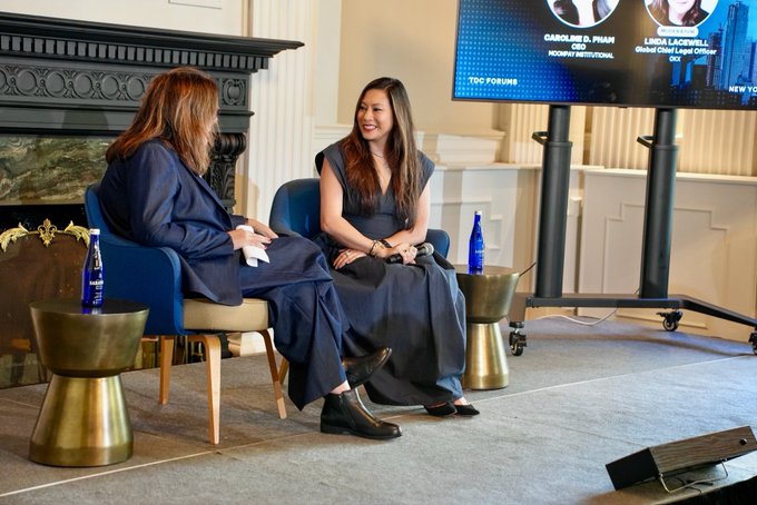 Two women sit on a small stage during a panel discussion, with one appearing engaged in conversation while the other listens attentively. A screen in the background displays the names "CAROLINE D. PHAM, CHAIR, CFTC, HODGEPARTY INSTITUTIONAL" and "LINDA LACEWELL, Global Chief Legal Officer," identifying them as speakers. The setup, featuring elegant gold side tables with bottled water and a fireplace backdrop, suggests a formal industry event—aligning with the post about a conversation involving Linda Lacewell and the Digital Chamber on clarity in crypto regulation.
