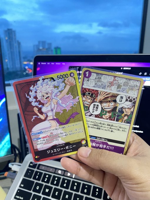 A hand holds two Japanese trading cards from the One Piece Card Game featuring the characters Jewelry Bonney and Monkey D. Luffy, with a laptop and city skyline visible in the background.
