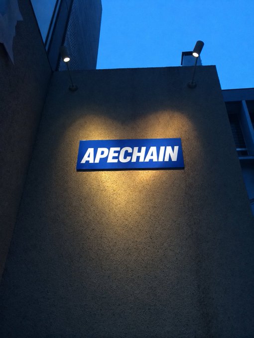 An illuminated blue APECHAIN sign mounted on a textured wall, lit by two spotlights at dusk.