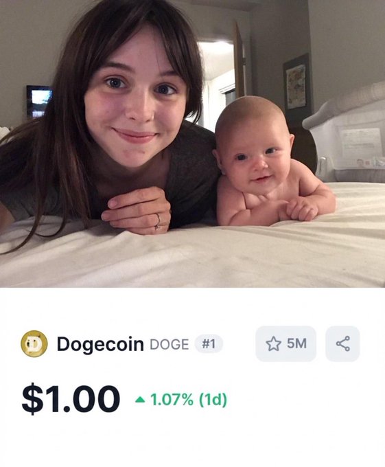 A selfie of a smiling young woman with long brown hair lying on a bed next to a baby propped on its arms, with a Dogecoin (DOGE) price widget below showing $1.00, up 1.07%.