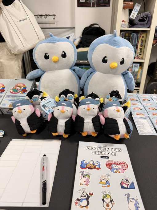 A display of Pudgy Penguins merchandise, including two large plush penguins, smaller plush toys in hoodies, and a 'Pudgy Penguins Singapore' sticker sheet.