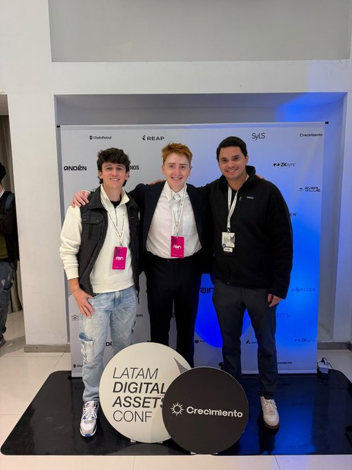 Three young men posing together at the LATAM Digital Assets Conf against a sponsor-branded backdrop, with company logos like ChainPlorer, REAP, and SVLS visible.