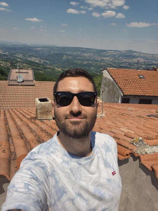A man wearing sunglasses and a white tie-dye t-shirt takes a selfie on a terracotta-tiled rooftop with a scenic, mountainous countryside view in the background, capturing a sunny Sunday moment that complements the cheerful "happy Sunday" sentiment of the post.