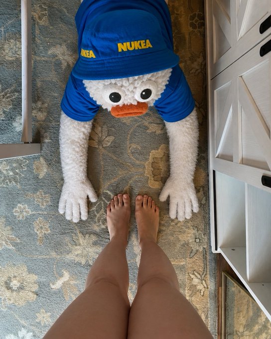 First-person view of someone's bare feet approaching a whimsical mascot plush toy dressed in blue 'NUKEA' apparel on a patterned carpet.