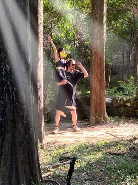 Casual outdoor photograph of a person in a forest setting wearing athletic shorts and a shirt, posing near trees with sunlight streaming through the branches.