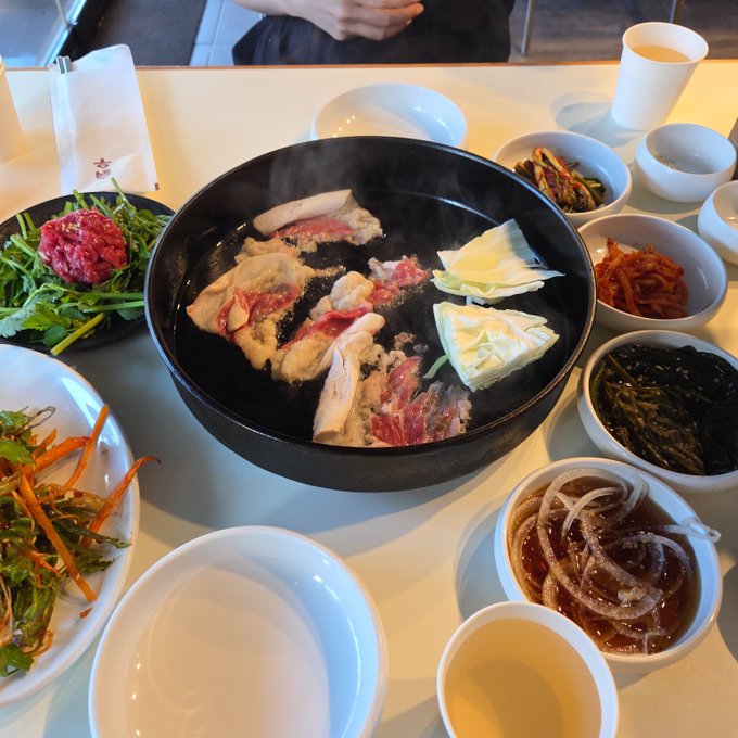 A Korean meal is spread on a table featuring a large black bowl of steaming bulgogi soup, surrounded by small side dishes including raw beef, vegetables, kimchi, and onions.