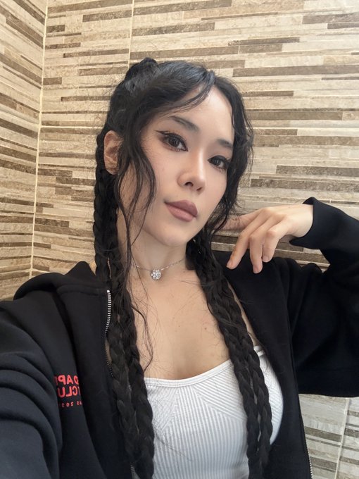 A selfie of a woman with long braided dark hair, wearing a black hoodie and a white top, posed against a textured stone wall background.