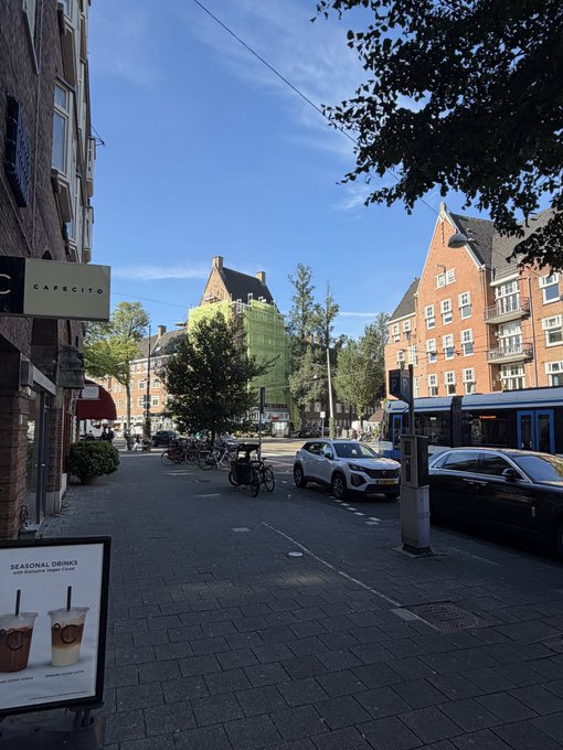 A sunlit Amsterdam street scene featuring a Capriccio café sign and seasonal drinks advertisement on the left, with brick buildings, parked cars, and bicycles lining the cobblestone road under a clear blue sky—capturing the bright, pleasant morning atmosphere referenced in the post.