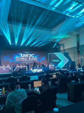 The image shows a large event hall with "UEX Global Alpha" and "Bitget" branding prominently displayed on screens and a lit stage, where attendees are seated at desks with laptops in a competitive trading tournament atmosphere. The accompanying post describes reporter Thành Phan live at the Bitget Trading UEX competition in Bali, Indonesia, humorously comparing the intense setup to a League of Legends arena.