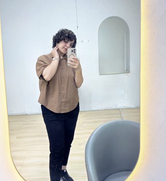 A person with short curly hair and glasses takes a mirror selfie in a sparsely furnished room, wearing a brown button-up shirt and black pants.
