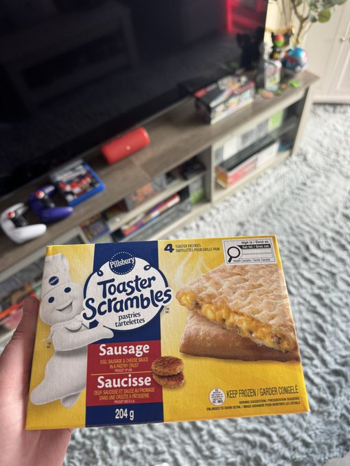 A hand holding a box of Pillsbury 'Toaster Scrambles' Sausage Pastry Tartlets in front of a TV stand with gaming consoles, books, and a plush rug.