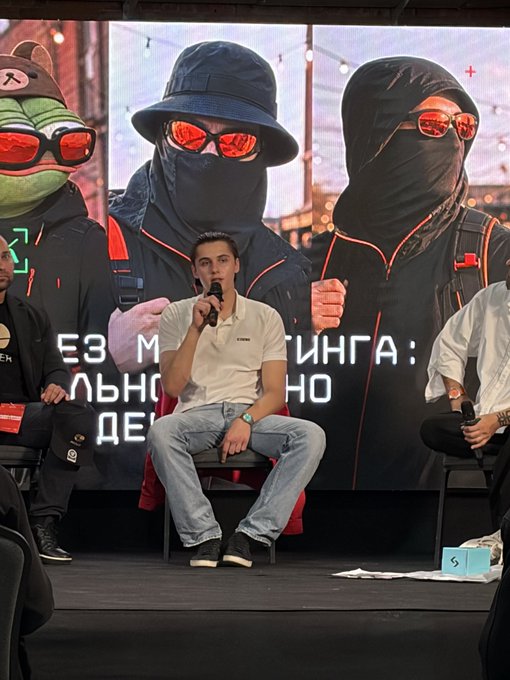 A man speaks into a microphone while seated on stage, with a backdrop featuring masked figures and partially visible Cyrillic text.