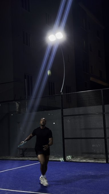 A nighttime photograph of a person on a blue padel/tennis court tossing a yellow ball in the air, illuminated by bright floodlights with a building visible in the background.