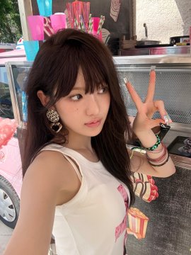 A young woman with dark hair makes a peace sign while wearing a white tank top and large earrings, posing in what appears to be a casual outdoor setting near a food stand.