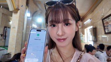 A young woman with sunglasses on her head takes a selfie showing a phone screen displaying a green checkmark, with other people visible in the brightly lit indoor background.