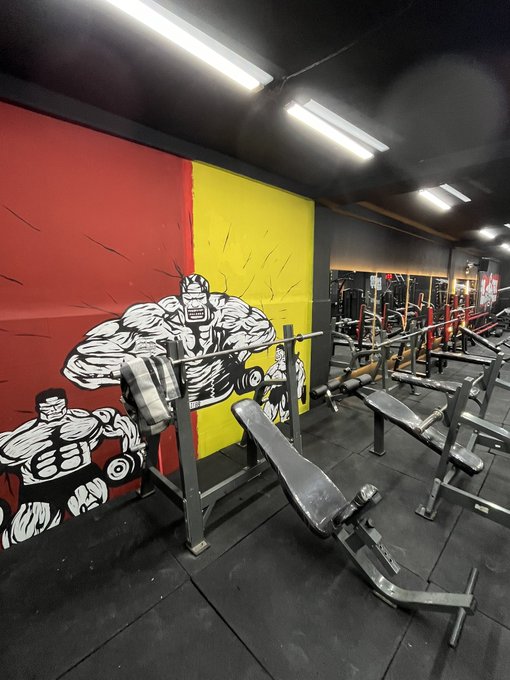 A gym interior features a bold wall mural of comic-style muscular figures lifting weights against red and yellow panels, alongside a bench press station. The motivational atmosphere aligns with the caption "Gn bros - Tomorrow we gonna try again," reflecting a determination to return and continue training after a tough session.