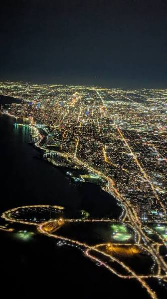 An aerial nighttime view of a coastal city illuminated with lights, showing streets, a harbor, and what appears to be a stadium or arena.