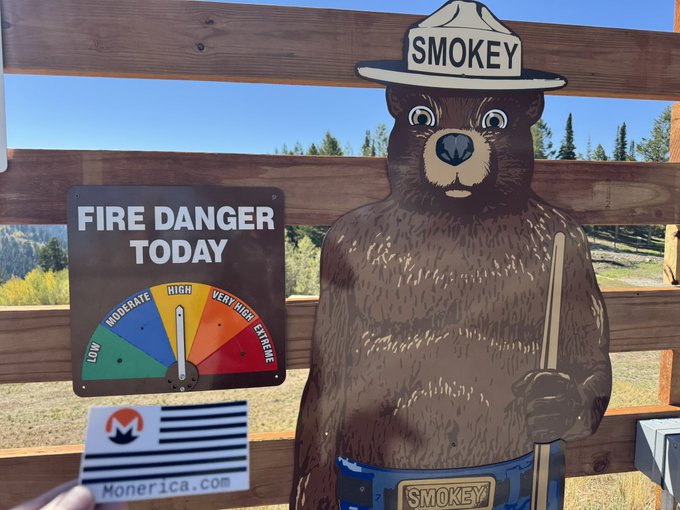 A cutout of Smokey Bear wearing his iconic ranger hat stands next to a 'Fire Danger Today' sign with the indicator pointing to 'High', with a Monerica.com card visible at the bottom.