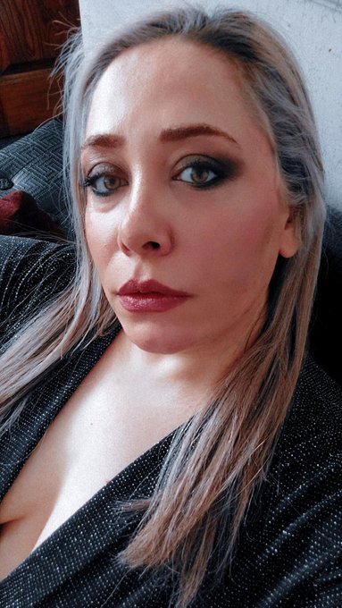 A close-up selfie of a woman with blonde hair, dark smoky eye makeup, and a black sparkly top, resting on what appears to be a couch.