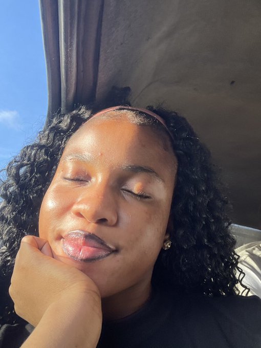 A close-up selfie of a woman with curly hair, her eyes closed, resting her chin on her hand under bright sunlight.