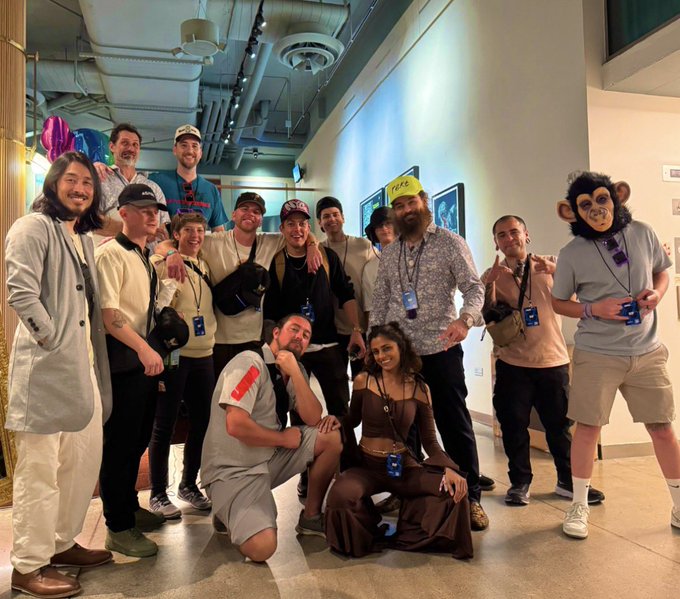 Group photo of approximately a dozen people posing together indoors, including someone wearing a large monkey head mask and another in a banana hat.