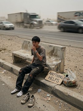 A young man sits on a weathered concrete bench by a dusty roadside with cars and trucks passing in the background, holding a cardboard sign that reads 'ANYTHING HELPS'.