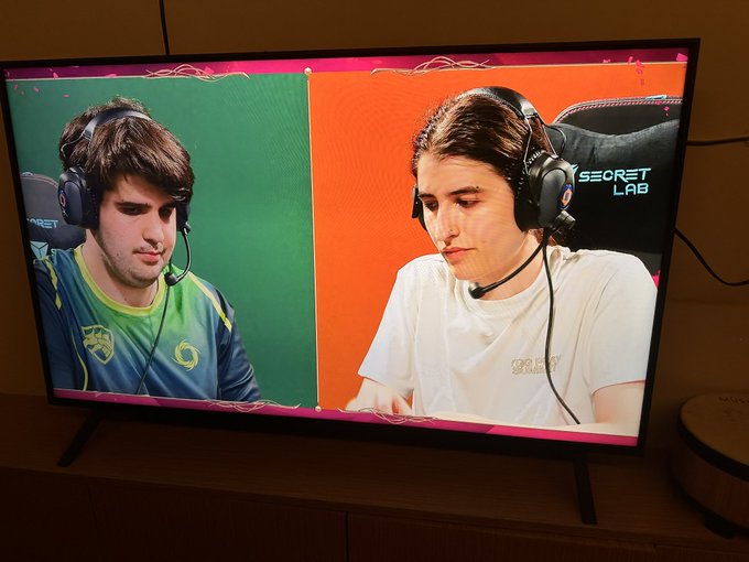 A TV screen shows two gamers wearing Secretlab headsets competing against each other, with one player on a green background wearing a green/yellow jersey and the other on an orange background in a white shirt. The "SECRET LAB" branding is visible on the headset, fitting the post's suggestion of forming a Riftbound gaming team sponsored by the chair brand.