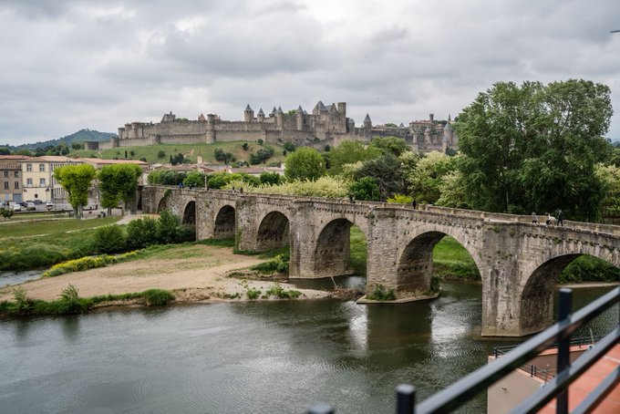 A stone arch bridge spans a river, with the historic fortified citadel of Carcassonne visible in the background under a cloudy sky.