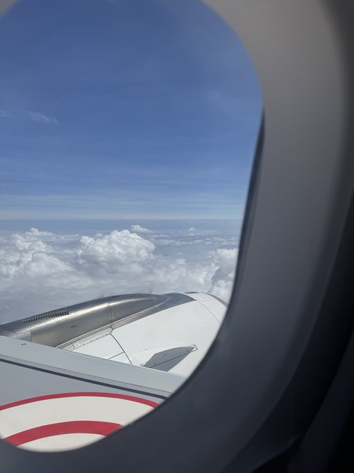 A view from an airplane window showing the wing and engine above white clouds and blue sky.