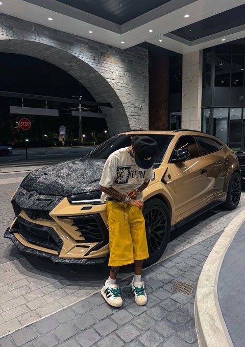 A person wearing a mask, a graphic t-shirt, yellow shorts, and white sneakers stands next to a customized gold-colored Lamborghini Urus SUV with a textured black hood, parked outside a building at night.