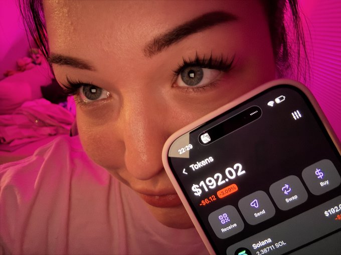 A close-up selfie of a woman with long lashes holding a smartphone displaying a crypto wallet with a token balance of '$192.02' and a Solana balance of '2.3571 SOL'.