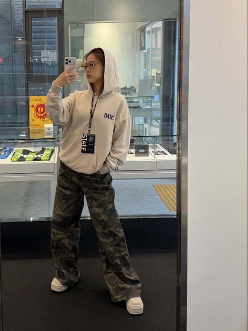 A person wearing a beige "BAYC" hoodie poses for a mirror selfie in what appears to be a retail or office space, paired with camouflage pants and chunky white sneakers. The image showcases the oversized, casual fit of the hoodie, aligning with the post's description of it as a cozy, boyfriend-style staple perfect for everyday wear.
