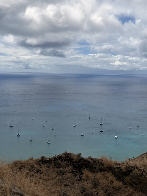 Anchored sailboats dot a calm bay under cloudy skies, viewed from an elevated coastal vantage point with dry brush in the foreground. The caption references a Solana address associated with "GM" (good morning), fitting the tranquil seaside scene.