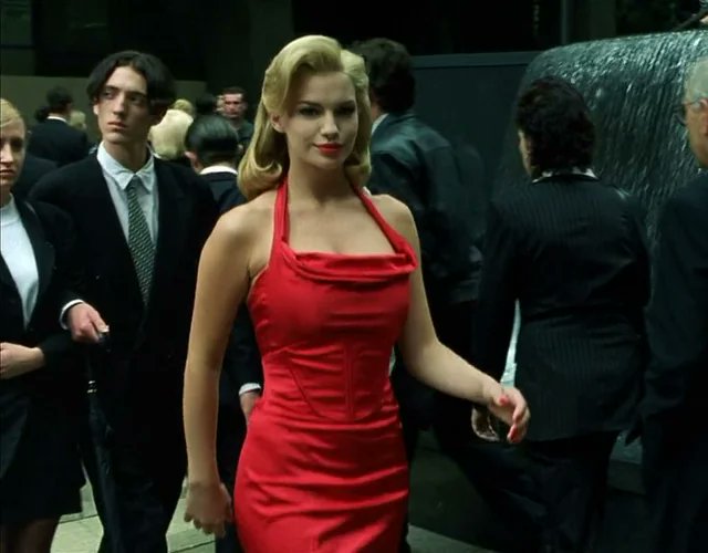 A blonde woman in a striking red halter-neck dress walks through a crowd of people in formal attire, also appearing to be a scene from a film.