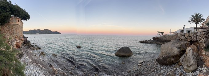 Coastal sunset scene with calm blue water, rocky shoreline in the foreground, small islands in the distance, and a building with a palm tree visible on the right side under a pale orange and blue sky.