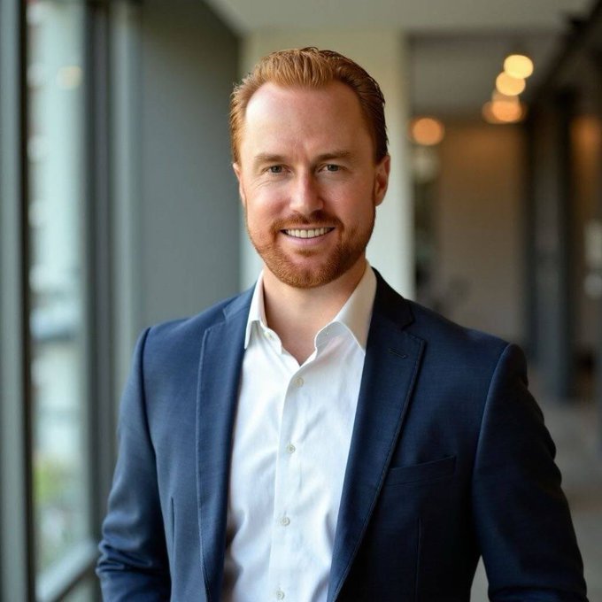 A smiling man with reddish hair and beard, wearing a navy blazer over a white dress shirt, stands in a modern office corridor with large windows. The image appears to be a professional headshot accompanying the announcement of his new role as Vice President of Sales.