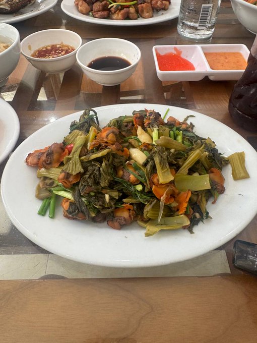 A plate of stir-fried greens with meat served on a wooden table alongside small bowls of dipping sauces and a plate of fried items.