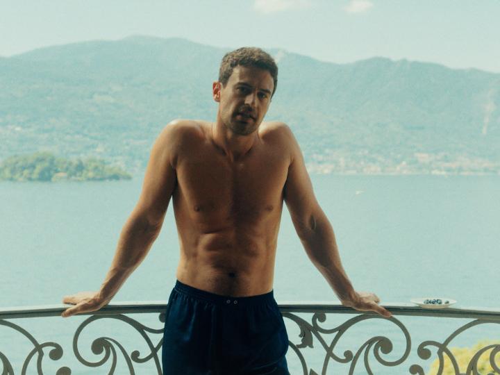 A shirtless, lean man with a beard stands on a balcony overlooking a scenic lake and mountains, wearing only navy blue shorts. The caption "congrats to whoever is bouncing on it" suggests someone is admiring his well-toned physique, likely as part of a humorous or flirtatious social media post.