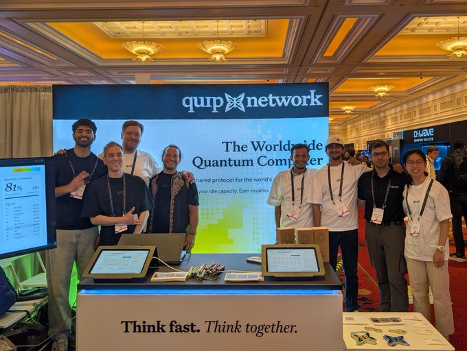 A group of seven people pose at the 'quip network' booth for 'The Worldwide Quantum Computer,' standing behind a counter with tablets and a sign reading 'Think fast. Think together.'