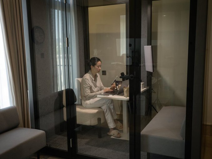 A woman in light-colored loungewear works on a laptop inside a glass-enclosed booth or study pod in a modern room.