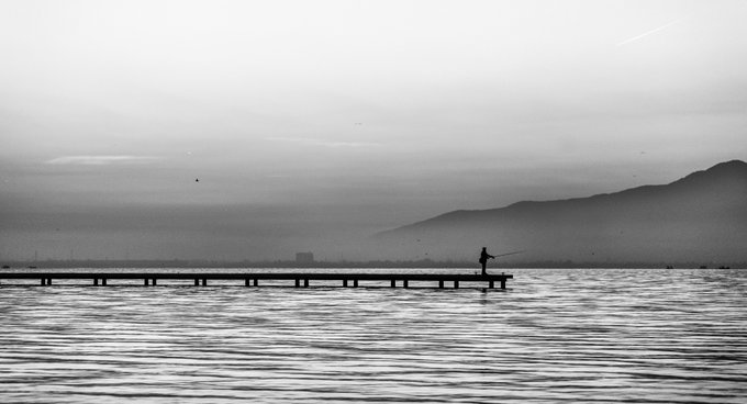 A lone fisherman stands on a long wooden pier extending into a misty, gray lake, with silhouetted mountains fading into the fog in the distance—evoking the melancholic mood of "My Own Oblivion" by Melih Ersahin, mirroring the artist's sentiment of fading into one's own oblivion.
