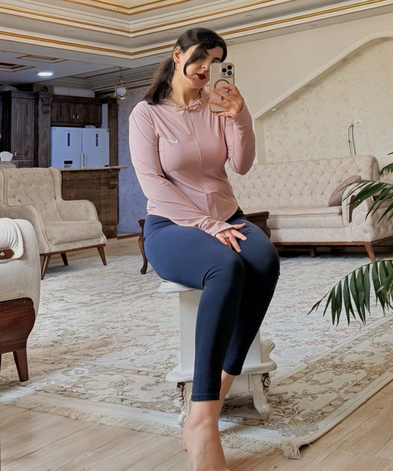 A woman takes a mirror selfie while sitting on a small white stool, wearing a pink long-sleeved top and dark blue leggings in a well-furnished living room.