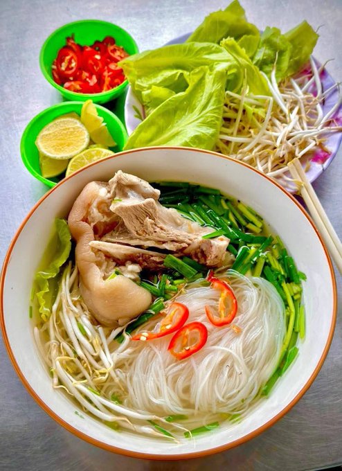 A large bowl of Vietnamese noodle soup containing broth, rice vermicelli, bean sprouts, greens, sliced red chili, and a large piece of bone-in pork, served with side plates of lime, chili, and lettuce.