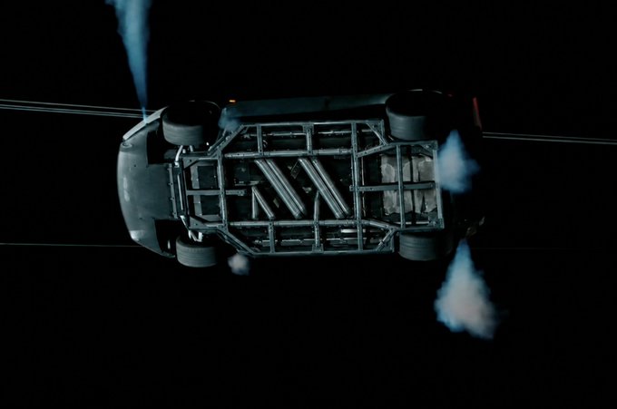 An overhead view of a dark vehicle's exposed chassis with smoke rising, appearing to be a scene from an action sequence.