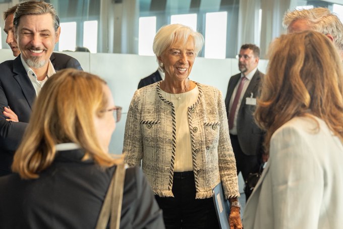 An older woman with short white hair, smiling and wearing a textured tweed jacket, stands at the center of a group of professionally dressed individuals in a modern, light-filled conference or institutional setting. The image likely depicts a formal European financial event, consistent with the post celebrating the launch of "Pontes," an ECB initiative for settling tokenised asset transactions in central bank money.