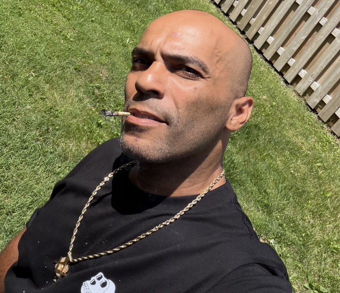 A bald man wearing a black t-shirt and gold chain lying on grass, holding a lit joint in his mouth.