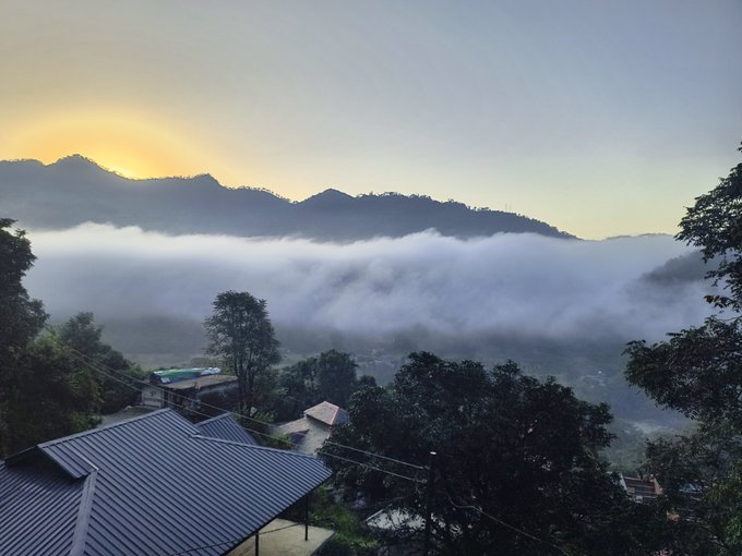 A scenic view of a misty mountain valley at sunrise or sunset, with rooftops and trees in the foreground.