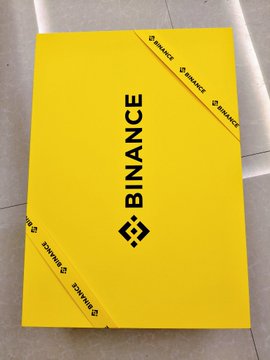 The image features a yellow box with a black "binance" logo, placed on a white surface. The box has been cut open, revealing its contents inside. The text "@binancezh 的网球礼盒", which translates to "@binancezh 的网球礼盒", is visible in the image as well.